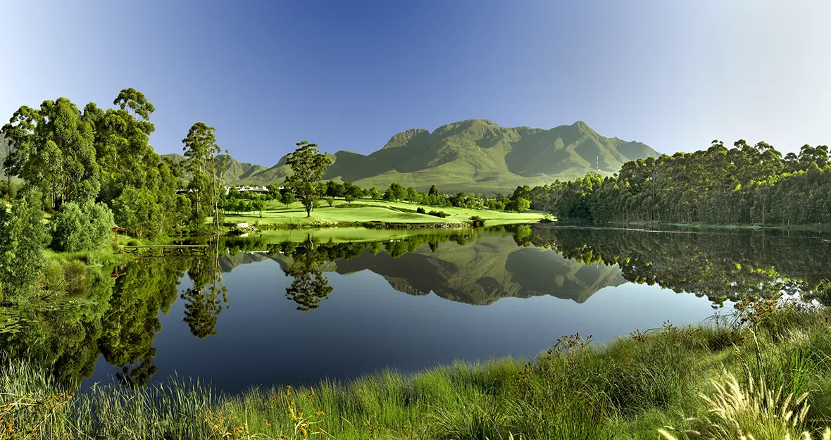 Eine ruhige Landschaft mit einem stillen See, in dem sich üppige grüne Bäume und ferne Berge unter einem klaren blauen Himmel spiegeln, umgeben von grasbewachsenen Ufern - das erinnert an die ruhigen Aussichten auf einer Golfreise in Südafrika.