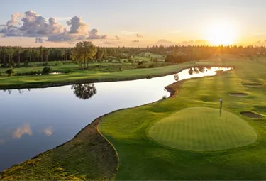 Ein malerischer Golfplatz bei Sonnenuntergang, perfekt für eine Golfrundreise durch Irland, mit einem Fluss, der sich entlang der grünen Fairways schlängelt, einem Putting Green mit einer Fahne, Sandbunkern und Bäumen im Hintergrund unter einem teilweise bewölkten Himmel.